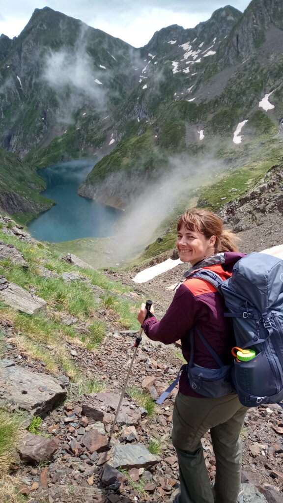 Me, standing with backpack, looking down over a mountain lake with snowy peaks in the background