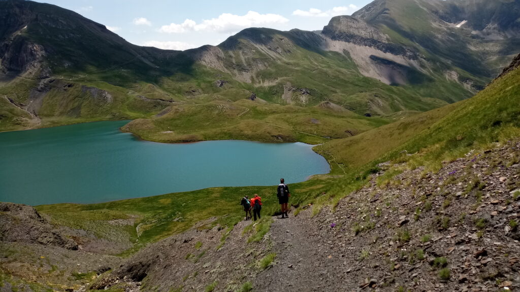 Three hikers seen from their back, descending down a rocky trail towards a clear bue mountain lake with grassy peaks in the background